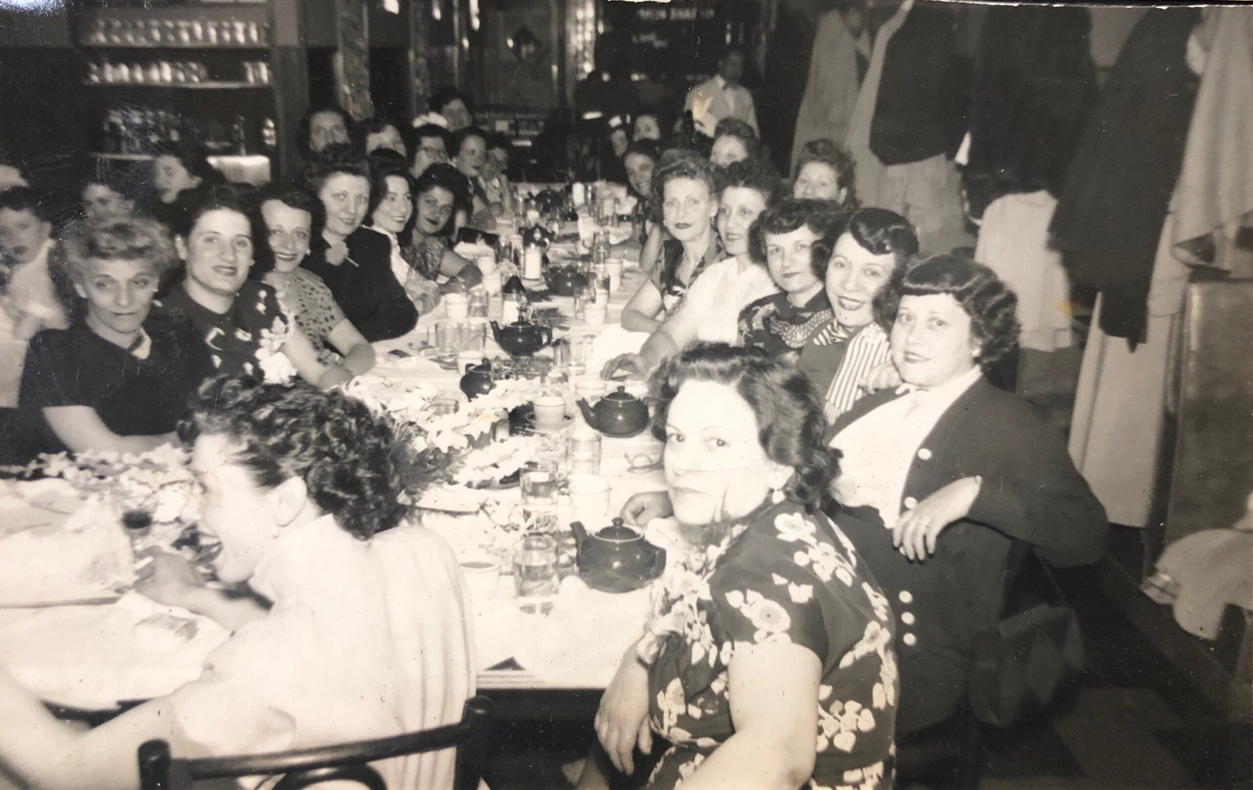 Classic black and white image of a number of women sitting around a long table, all facing the camera smiling and celebrating
