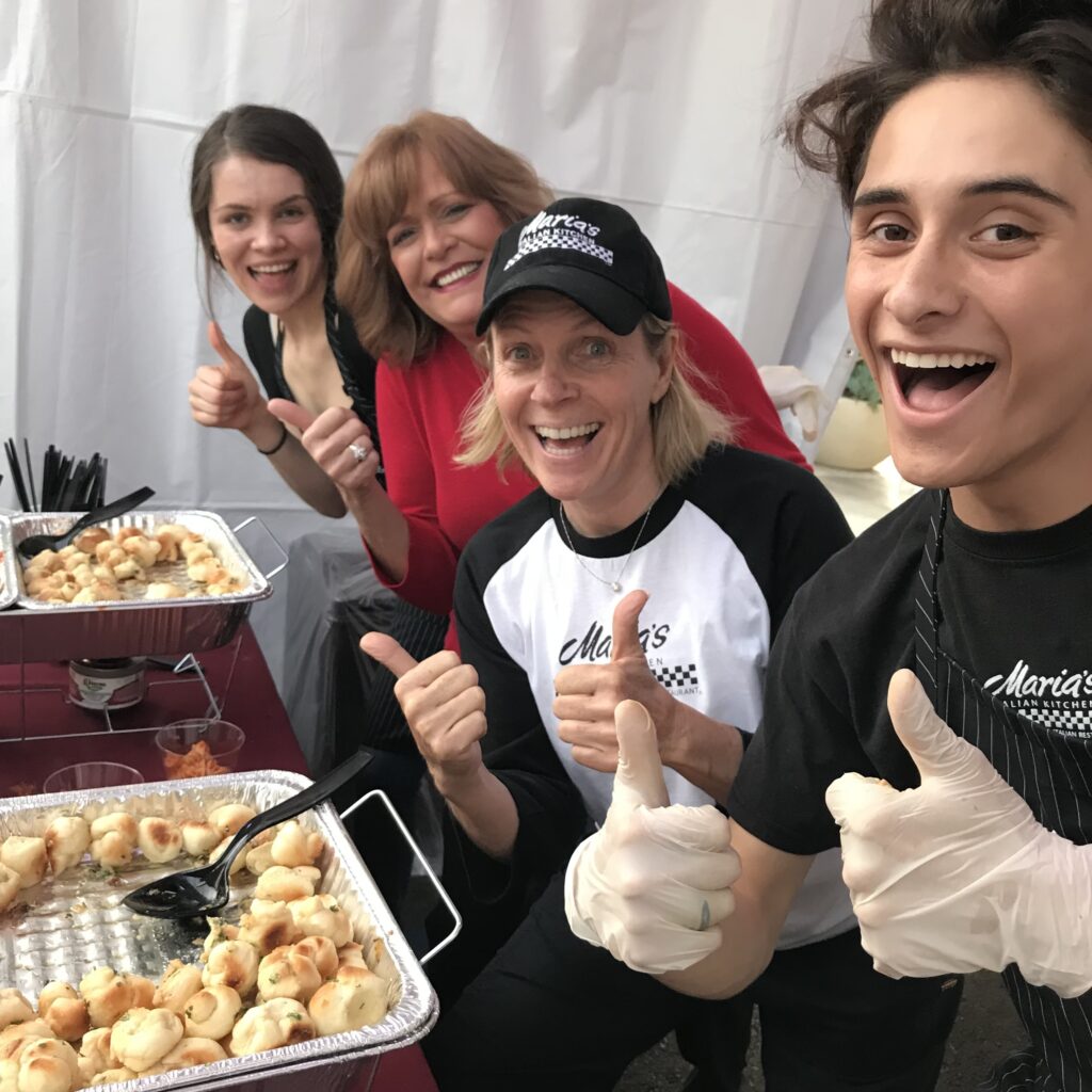 Maddy with 3 employees serving food from catering trays, all giving a thumbs up to the camera