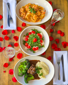 Bird's eye view of shrimp pasta, caprese salad, and short rib, surrounded by rose petals