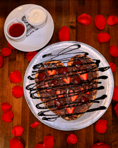 Top down picture of Nutella pizza on a plate with a small plate of cannoli cream and strawberry sauce above it to the left. Both plates are on a wooden table top, surrounded by rose petals